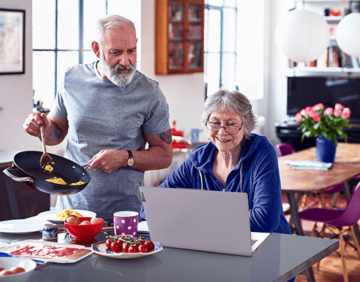 A older man cooking eggs and an older woman sitting at a counter top with a Lenovo IdeaPad.