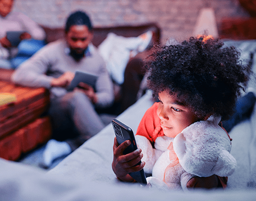 A child laying on the couch holding a stuffed animal and looking at their Lenovo ThinkPhone with someone faintly pictured in the background also looking at their device.