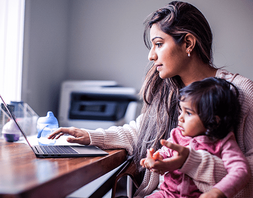 A woman sitting in a chair at a desk holding her child with one hand whilst scrolling on her Lenovo IdeaPad with the other.
