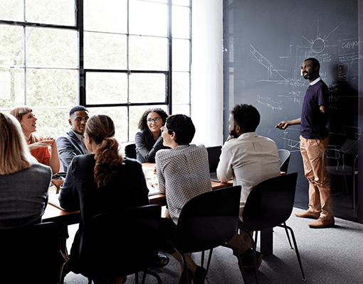 A group of people seated at a table during a meeting, with one person standing at a chalkboard.